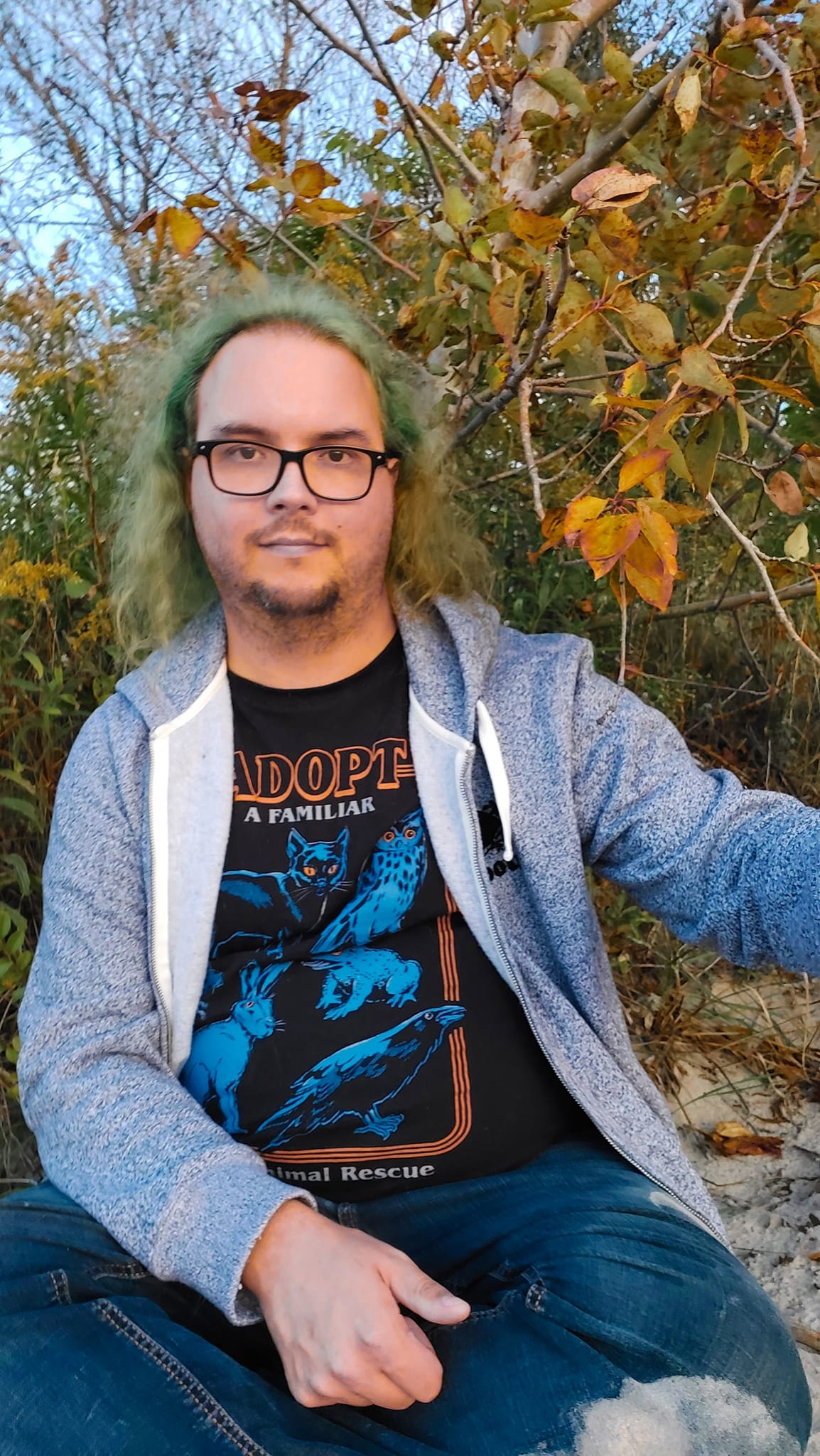 A man with green hair wearing a swetter sits on the soft white sand in front of a shrub which has lost most of its leaves